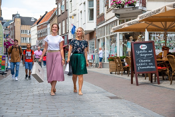 Twee vrouwen lopen met winkelzakken door de binnenstad van Roermond, langs winkels en een terras met menubord.