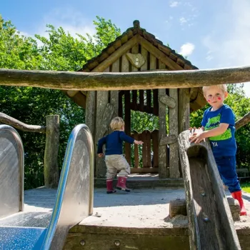 Twee jonge kinderen spelen bij een houten speelhuisje met glijbaan in een groene buitenspeeltuin in Limburg.