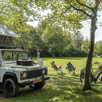 Twee kampeerders ontspannen in het gras naast hun camper tijdens het kamperen in Limburg, met uitzicht op een groene open plek.