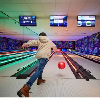 Kind gooit een bowlingbal in een kleurrijke hyperbowlingbaan bij de Leistert, met verlichte banen en schermen op de achtergrond.